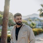 Bearded man with hands in pockets posing outdoors. Stylish outfit in natural setting.
