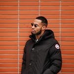 Stylish adult man wearing sunglasses and winter coat, leaning against a brick wall.
